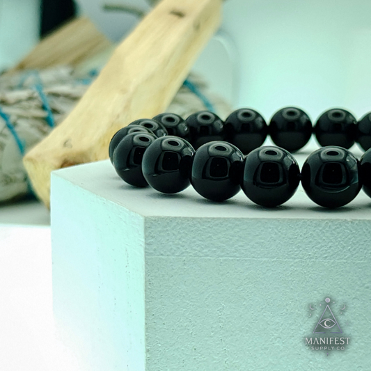 Black beaded bracelet on a light blue block with Manifest brand logo.