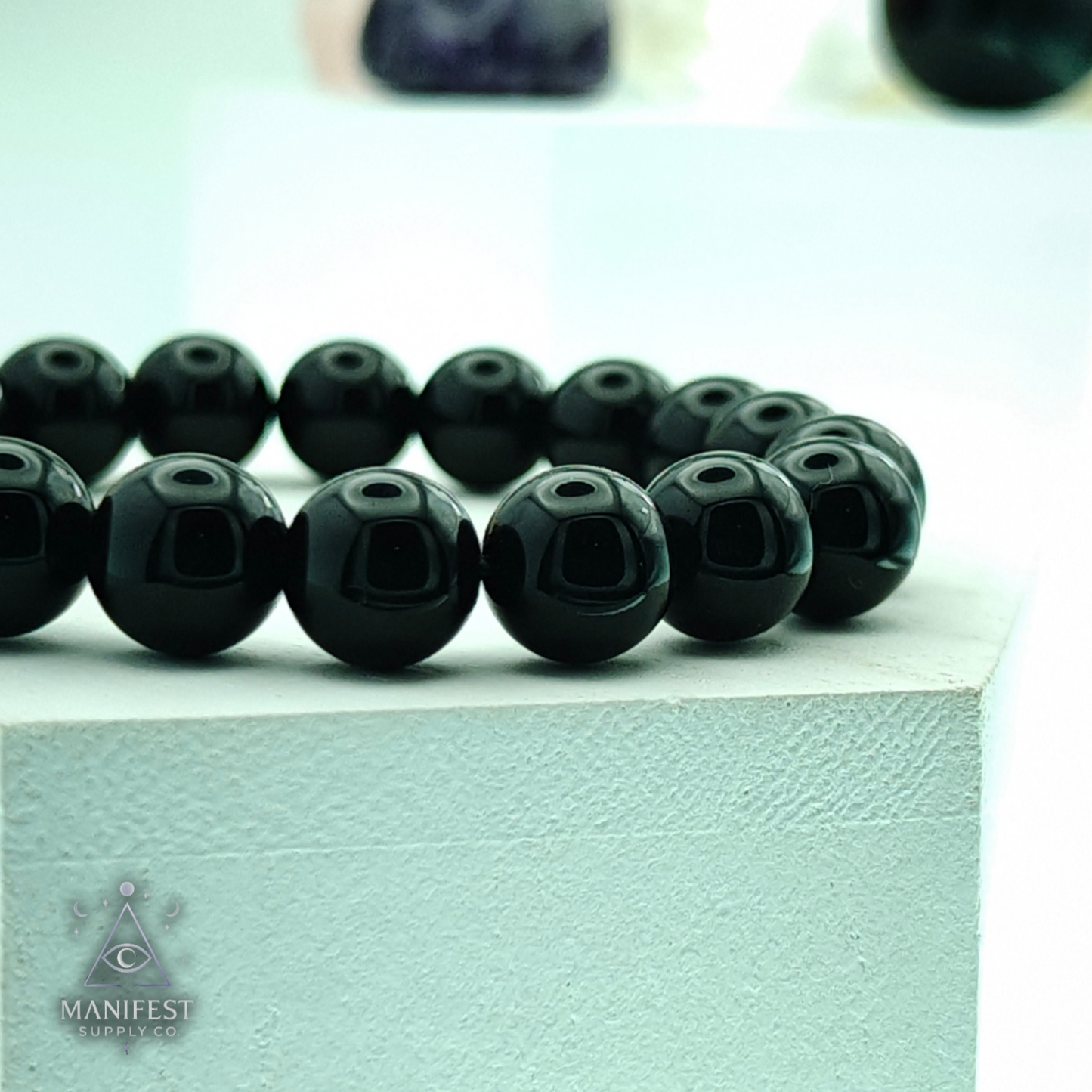 Black beaded bracelet on a light green surface with 'Manifest' branding.