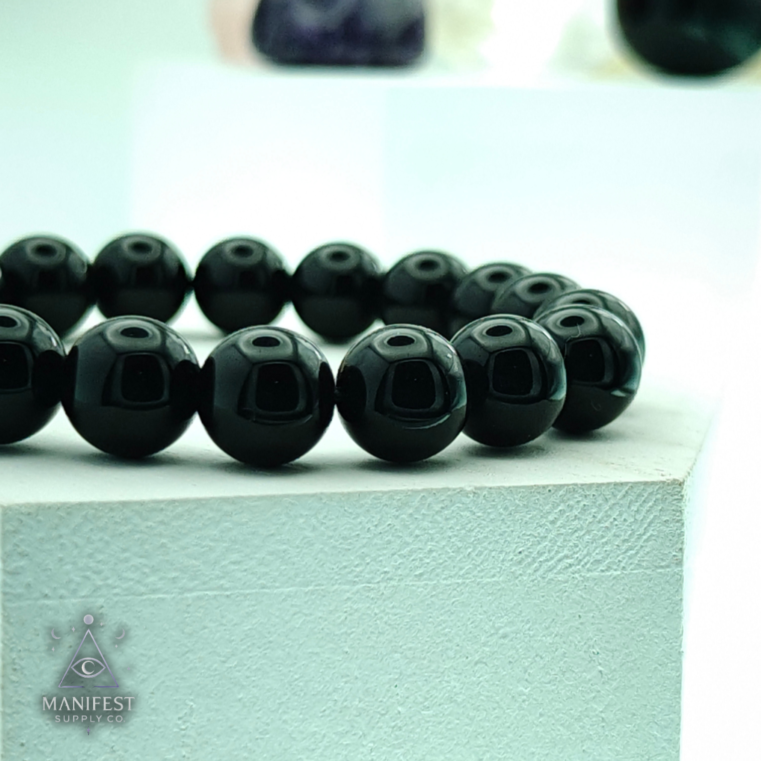 Black beaded bracelet on a light green surface with 'Manifest' branding.