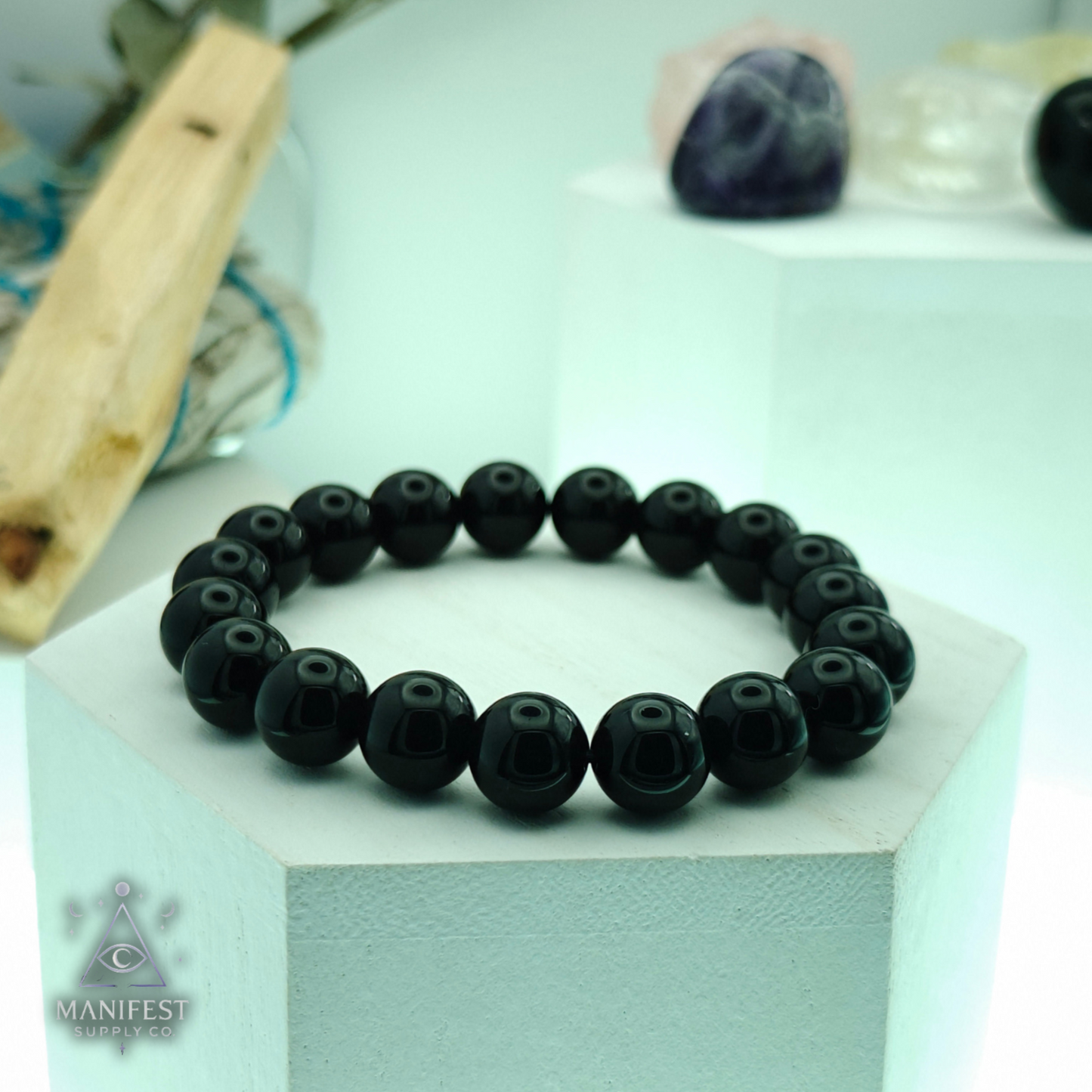 Black beaded bracelet on a white box with 'Manifest' branding, on a light green background.