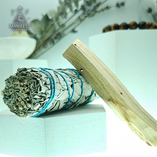Rolled sage smudge stick with wooden handle on a light surface, Manifest Supply Co. branding visible.