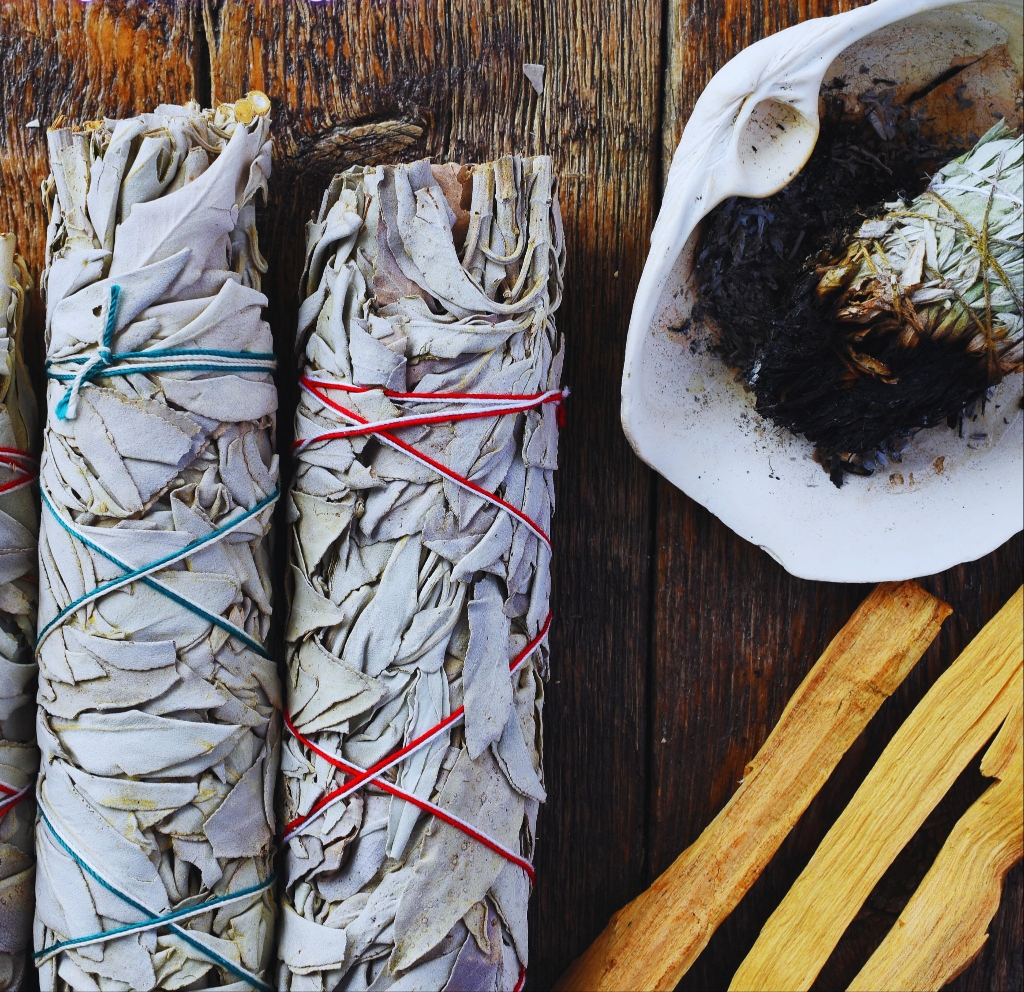 Two smudge sticks tied with colorful strings on a wooden surface with a shell and burning sage.